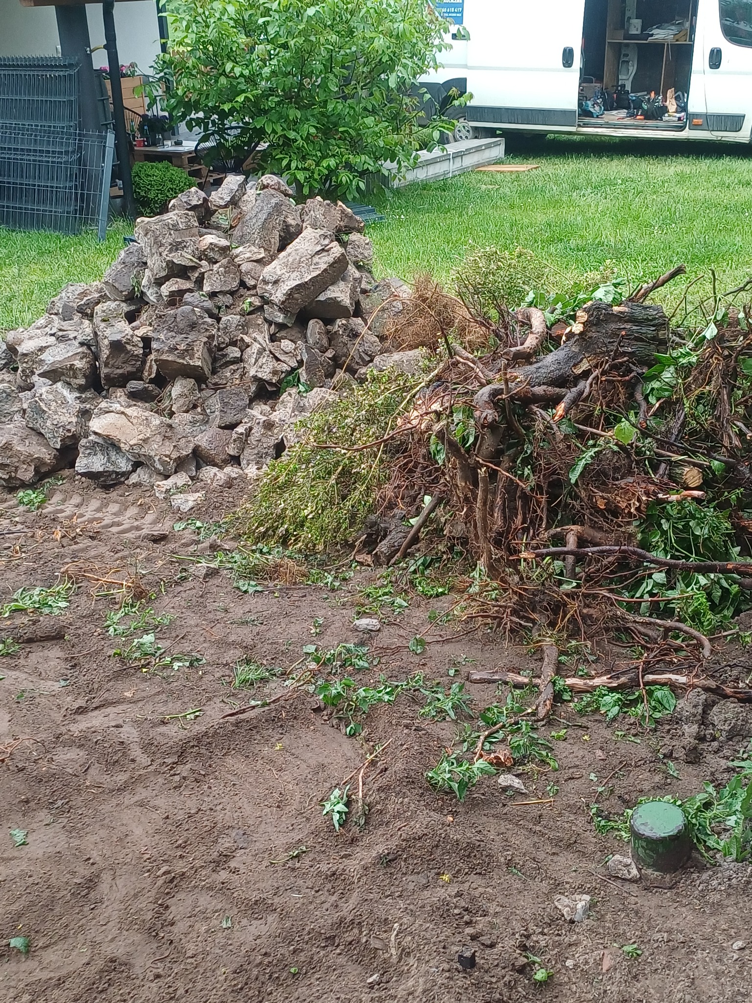 Góra kamieni i wykopane korzenie drzew na błotnistym terenie z widocznymi śladami opon, prawdopodobnie po niwelacji terenu. W tle zaparkowany biały bus.