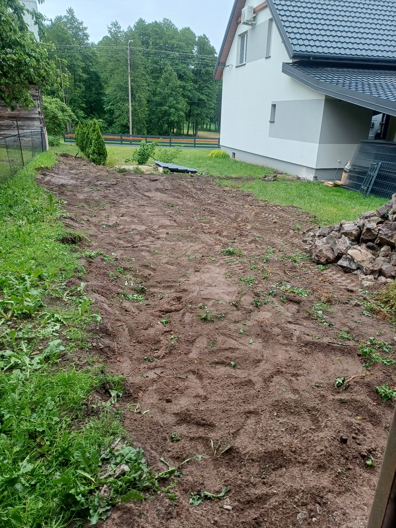 Przygotowany teren pod budowę domu jednorodzinnego z widocznymi śladami opon i stertą kamieni. Częściowo usunięta darń, widoczny budynek w tle.