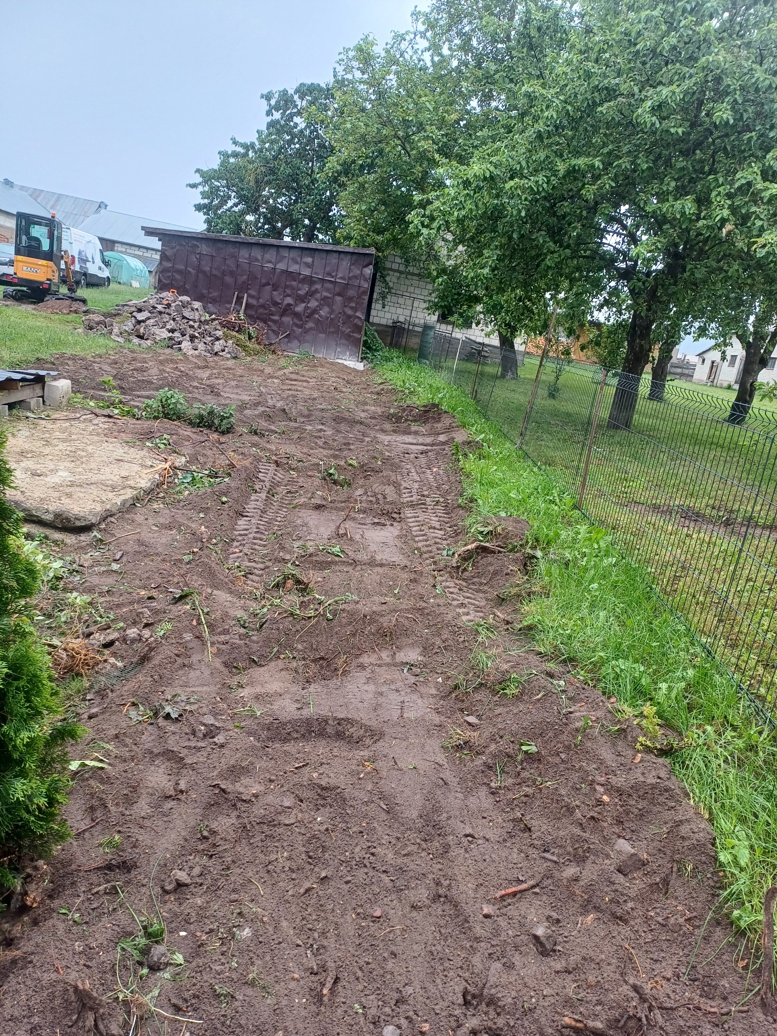 Przygotowany teren pod budowę z widocznymi śladami opon i niewielkim wykopem. W tle widoczny mały traktor i budynek gospodarczy.