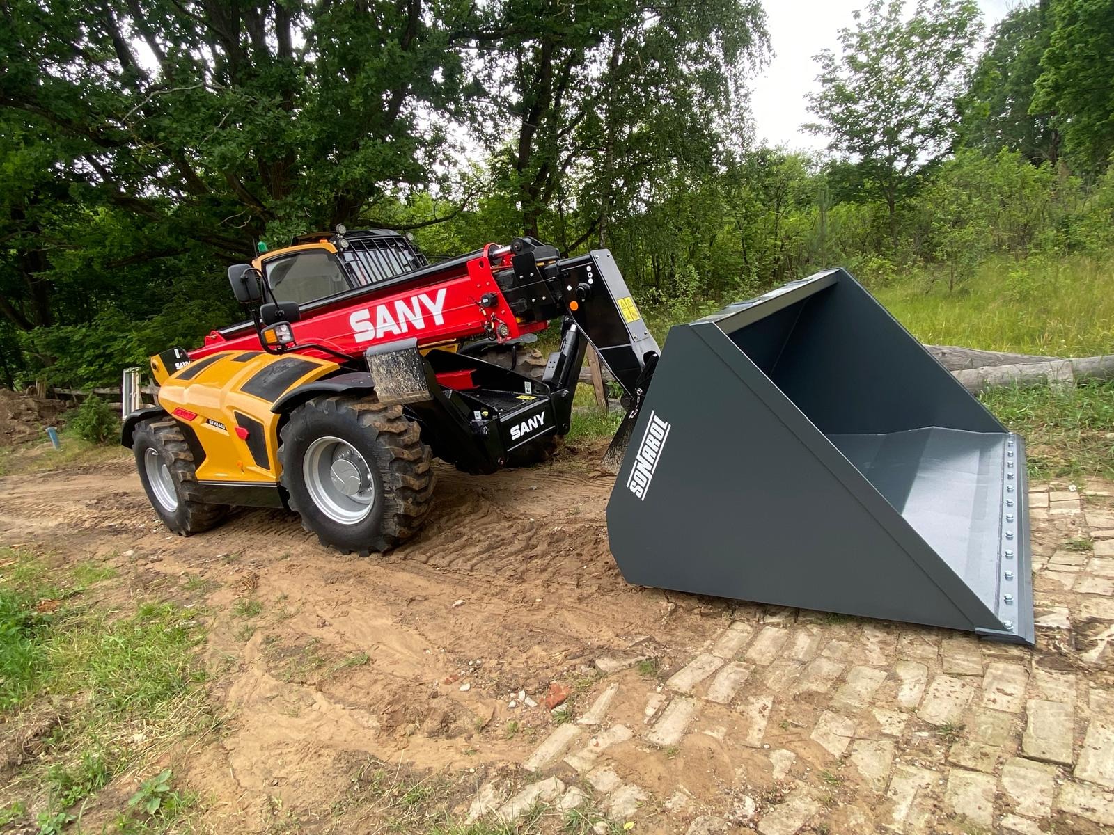 Żółto-czerwona ładowarka teleskopowa SANY z szarą łyżką SONAROL na tle zieleni. Maszyna stoi na ubitej ziemi z elementami kostki brukowej.