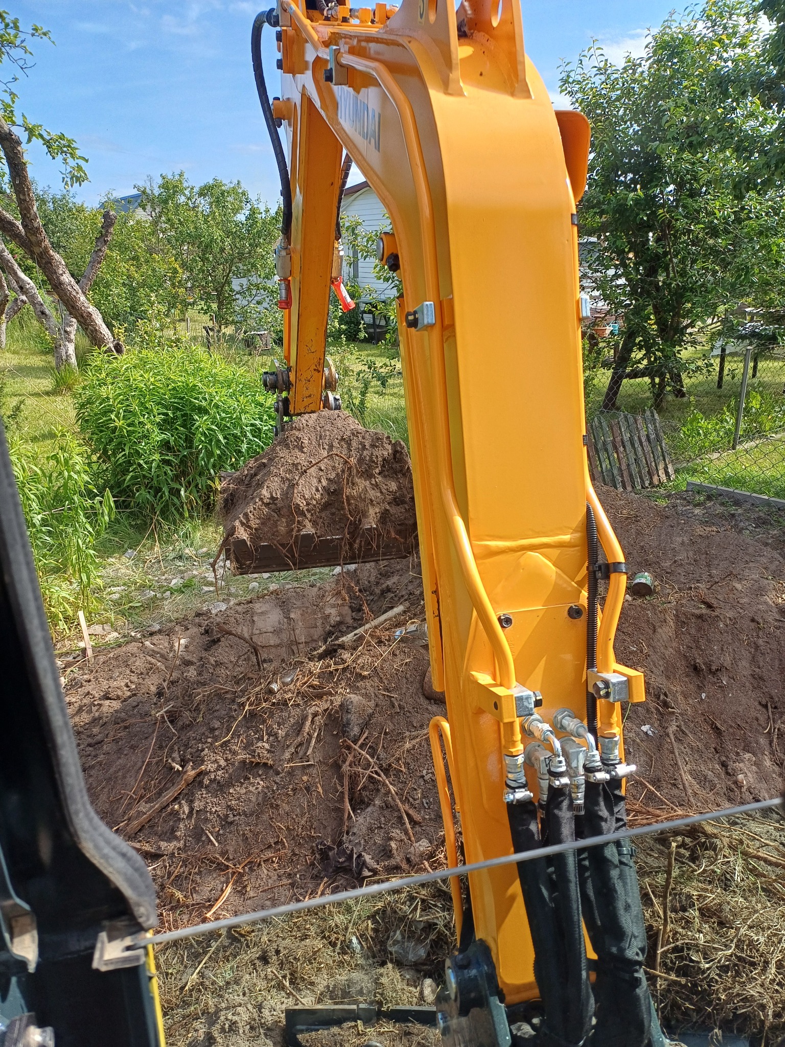Żółta koparka Hyundai w trakcie prac ziemnych w Białymstoku, z bliska widoczny nabierak z ziemią i korzeniami, w tle zieleń ogrodu.