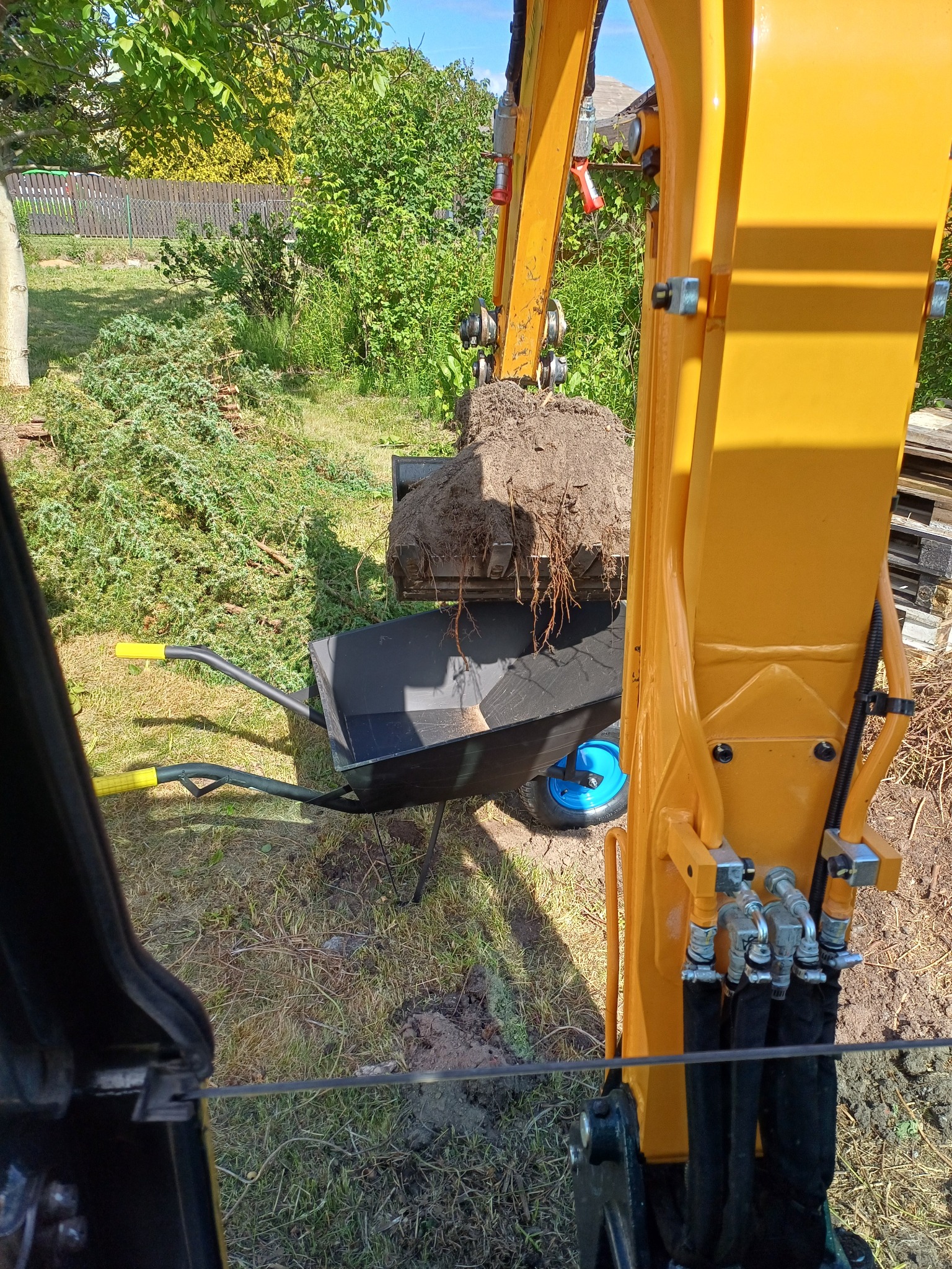 Żółta koparka z łyżką pełną ziemi, którą opróżnia do taczki. Widok z kabiny operatora na prace ziemne w ogrodzie.