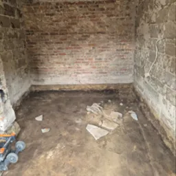 Kanciapa przed remontem: surowe ściany z cegły i betonu, widoczne ślady starej farby i gruz na podłodze. W rogu pomieszczenia stoi narzędzie budowlane.