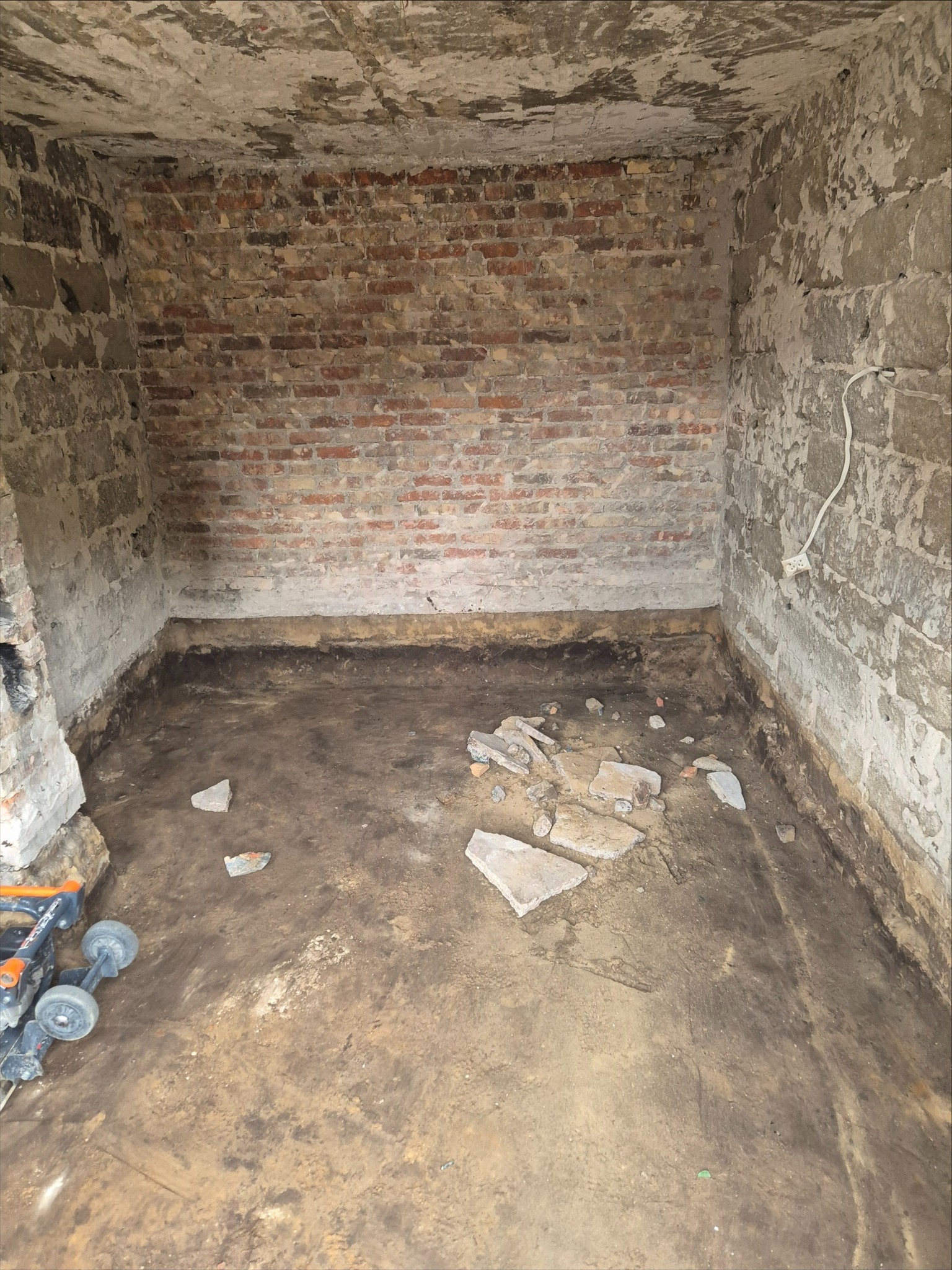 Kanciapa przed remontem: surowe ściany z cegły i betonu, widoczne ślady starej farby i gruz na podłodze. W rogu pomieszczenia stoi narzędzie budowlane.