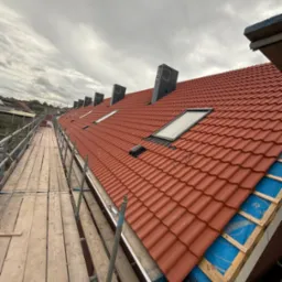 Czerwony dach z dachówką karpiówką, widok z rusztowania. Okna dachowe i kominy wentylacyjne na tle pochmurnego nieba. Fragment izolacji i membrany dachowej.