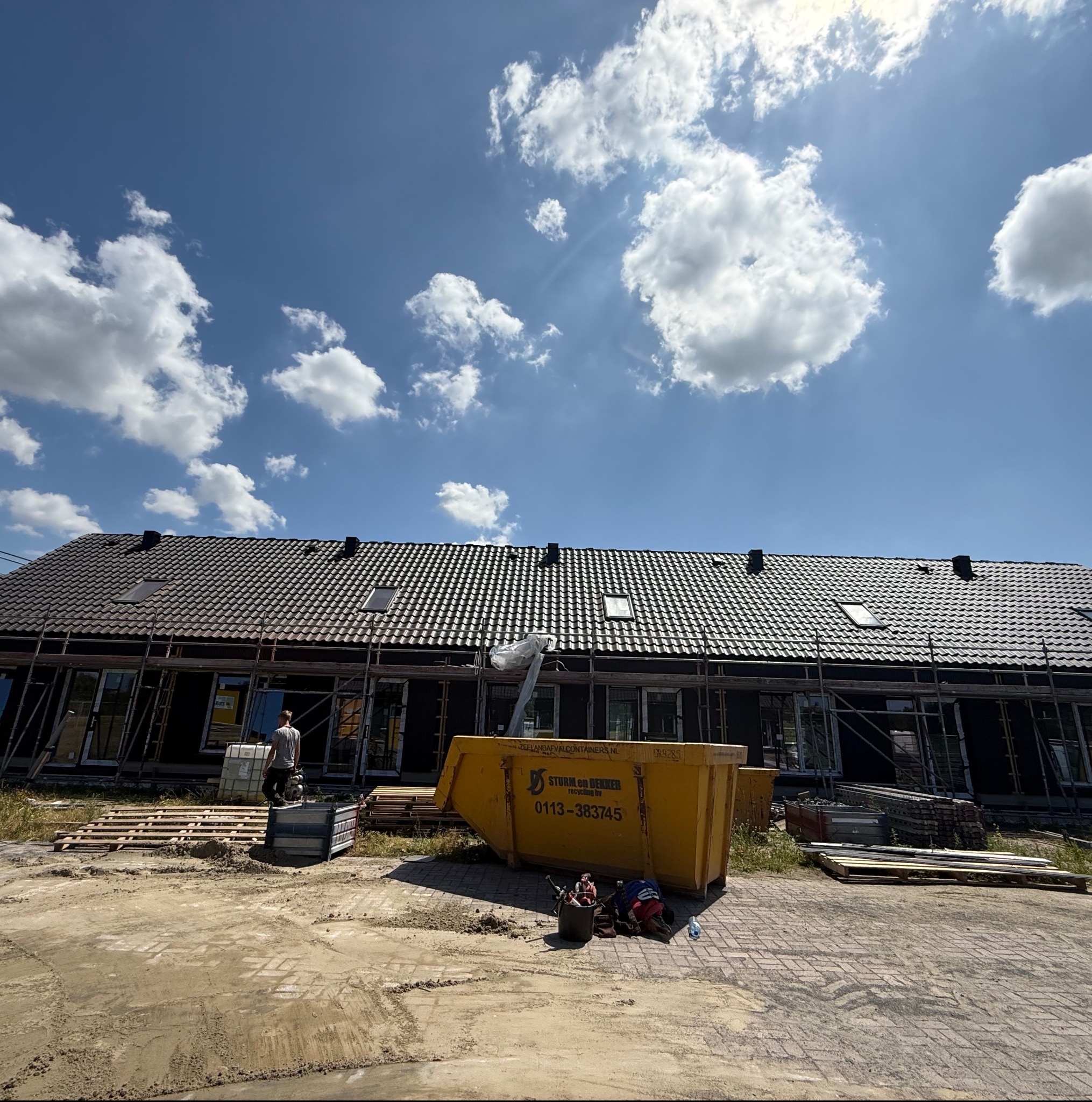 Nowe domy w zabudowie szeregowej w trakcie prac dekarskich. Widoczne rusztowania, kontener na odpady i materiały budowlane. Słoneczny dzień, błękitne niebo z chmurami.
