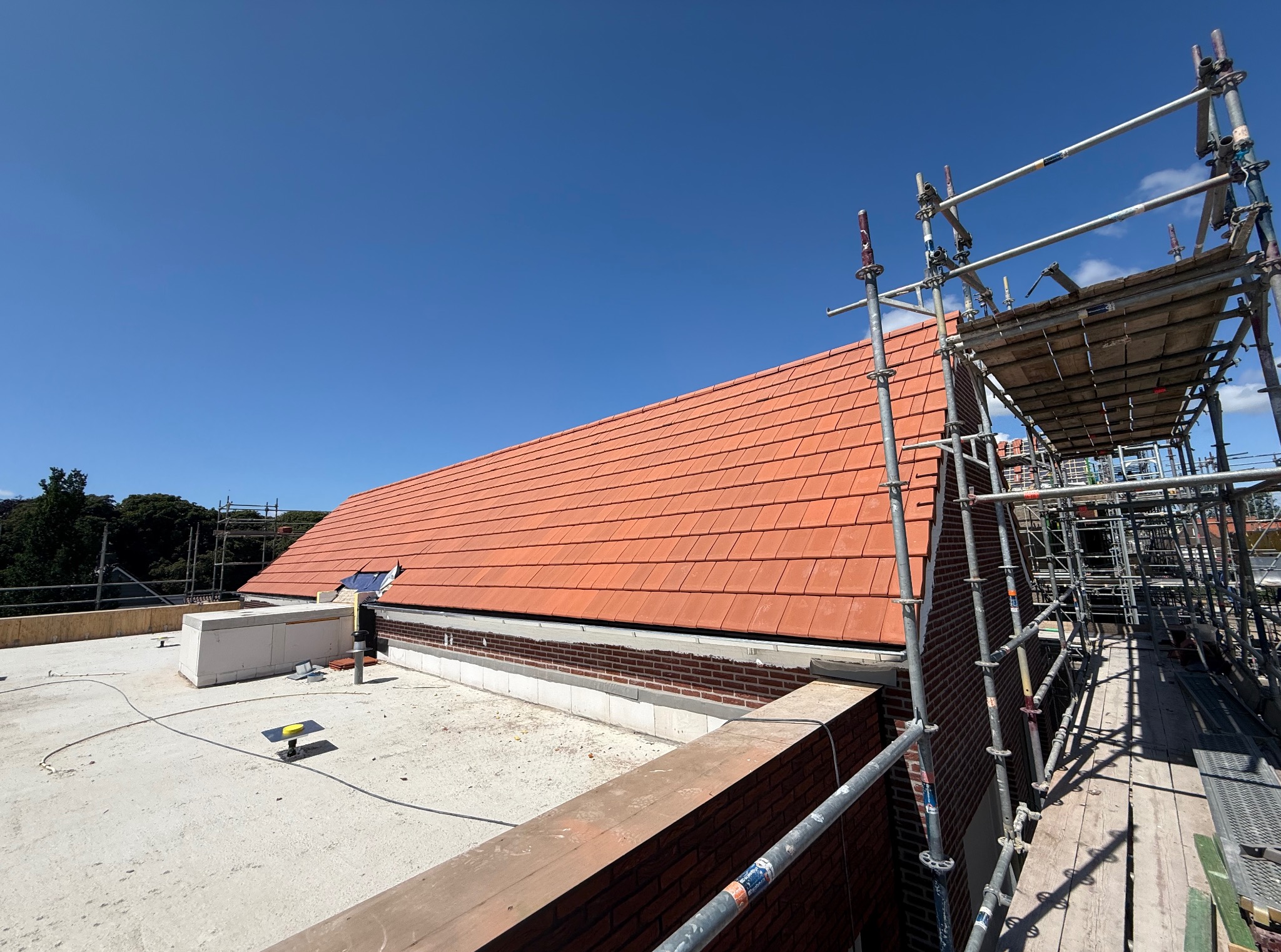 Czerwona dachówka na budynku w trakcie budowy, widoczna z rusztowania w słoneczny dzień. Częściowo ułożony dach i elementy konstrukcyjne.