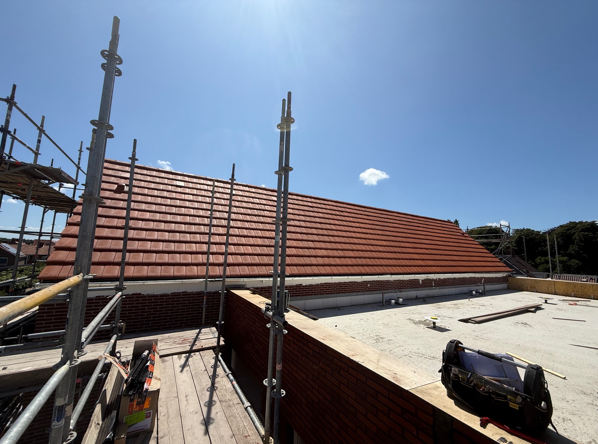 Czerwona dachówka na dachu domu, widok z rusztowania w słoneczny dzień. Częściowo widoczna ceglana ściana i narzędzia dekarskie.