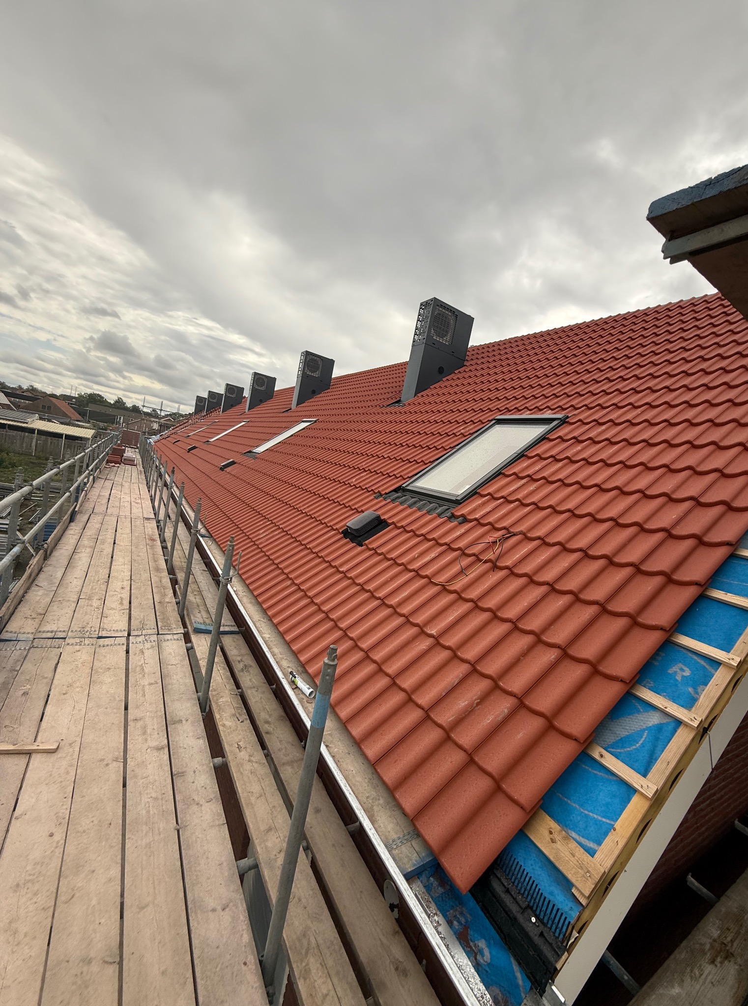 Czerwony dach z dachówką karpiówką, widok z rusztowania. Okna dachowe i kominy wentylacyjne na tle pochmurnego nieba. Fragment izolacji i membrany dachowej.