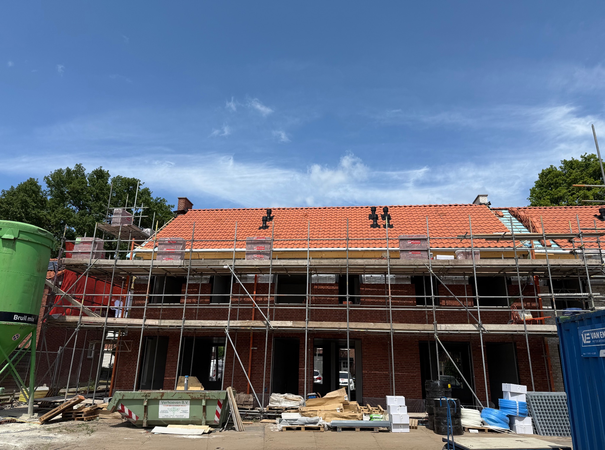 Budowa domu z czerwonej cegły, częściowo pokryty czerwoną dachówką, otoczony rusztowaniem. Widoczny kontener i materiały budowlane na placu budowy.