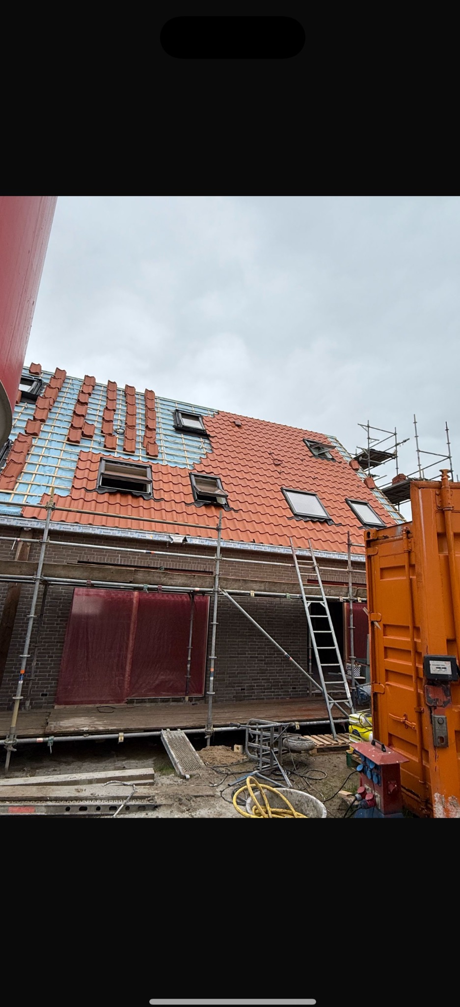 Częściowo pokryty czerwoną dachówką dach z oknami połaciowymi, widoczne rusztowania i kontener na placu budowy. Etap prac dekarskich w pochmurny dzień.