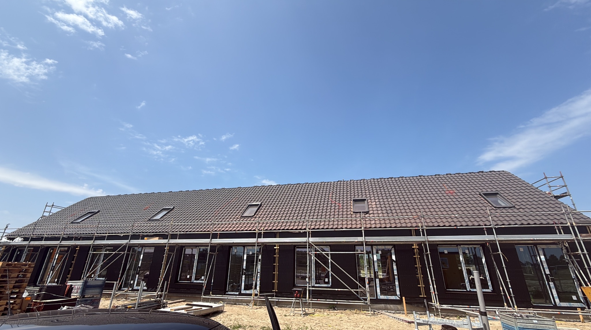 Budowa domu jednorodzinnego z ciemną dachówką i oknami dachowymi, otoczonego rusztowaniami na tle błękitnego nieba z chmurami.