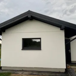 Nowoczesny, parterowy budynek z czarnym dachem i rynnami. Widoczny fragment elewacji, okno z czarną ramą i drzwi wejściowe. Tło stanowi pochmurne niebo i zieleń.