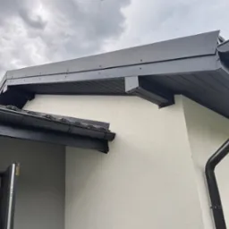 Detal czarnego dachu z rynnami i obróbkami blacharskimi na tle pochmurnego nieba. Nowoczesne wykończenie domu z widocznym fragmentem elewacji i okna.