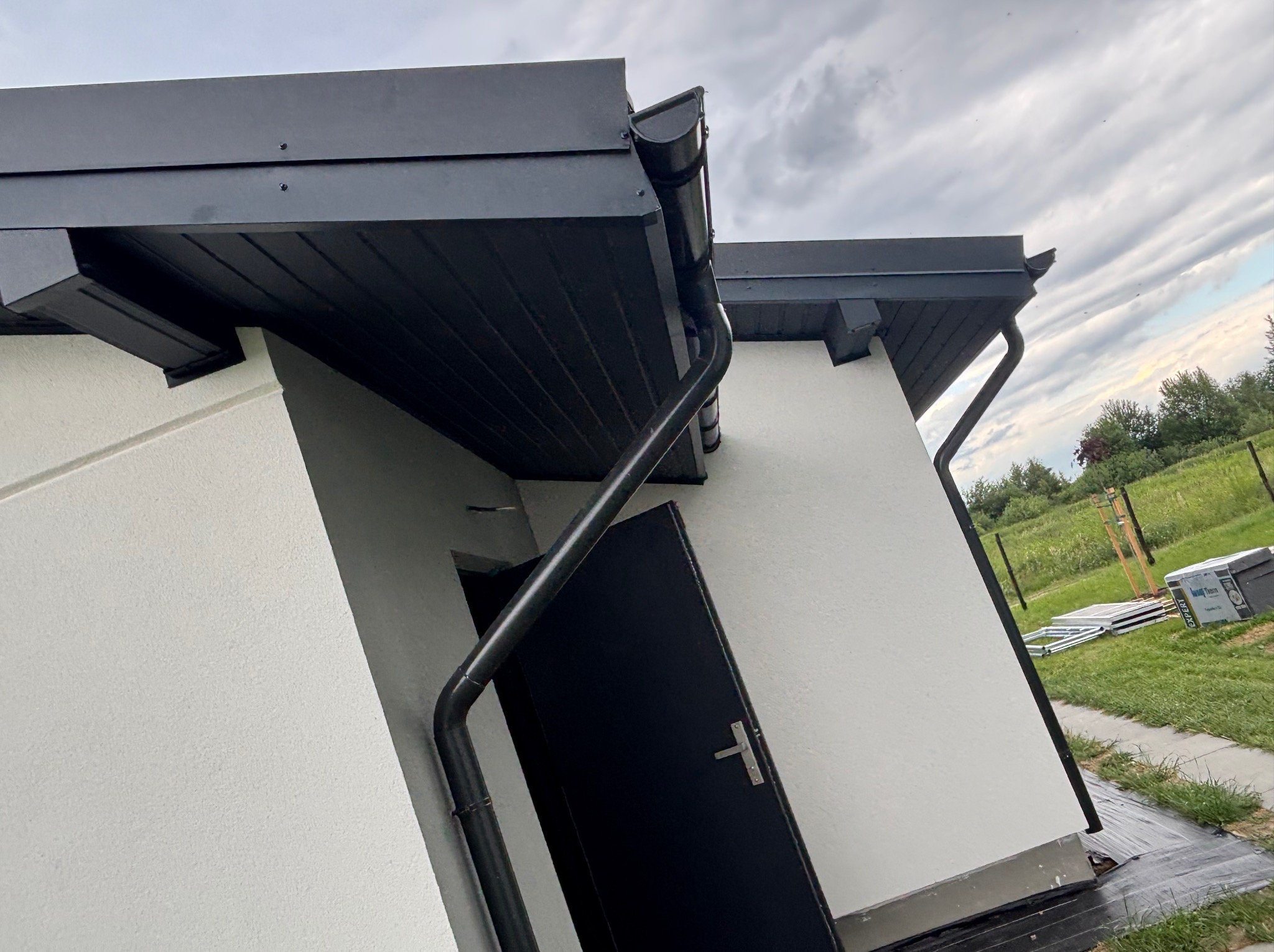 Nowoczesny budynek z czarnym dachem i rynnami, białą elewacją. Widoczne elementy wykończenia, kąt ujęcia z dołu podkreśla geometrię. W tle zieleń i pochmurne niebo.