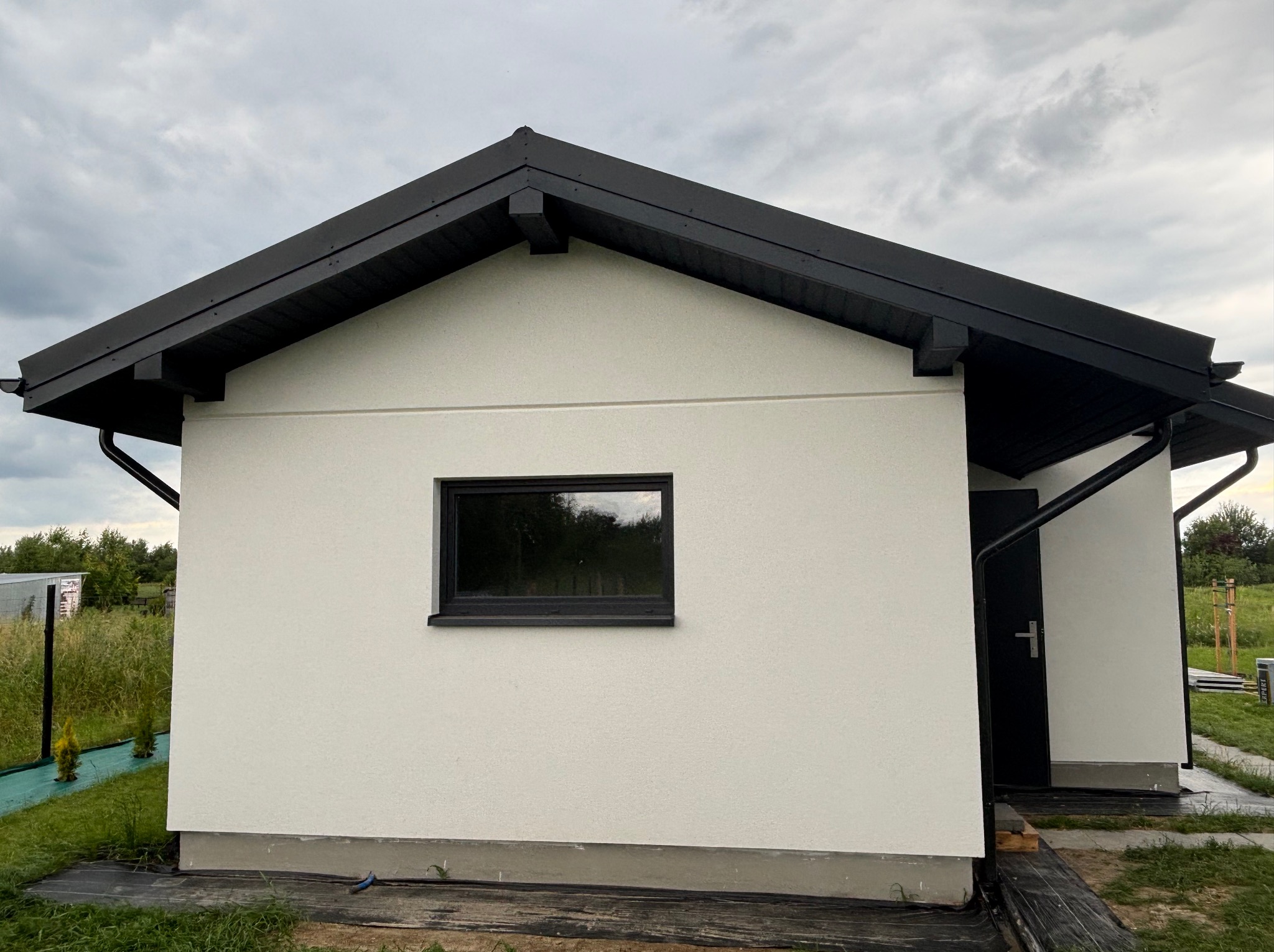 Nowoczesny, parterowy budynek z czarnym dachem i rynnami. Widoczny fragment elewacji, okno z czarną ramą i drzwi wejściowe. Tło stanowi pochmurne niebo i zieleń.