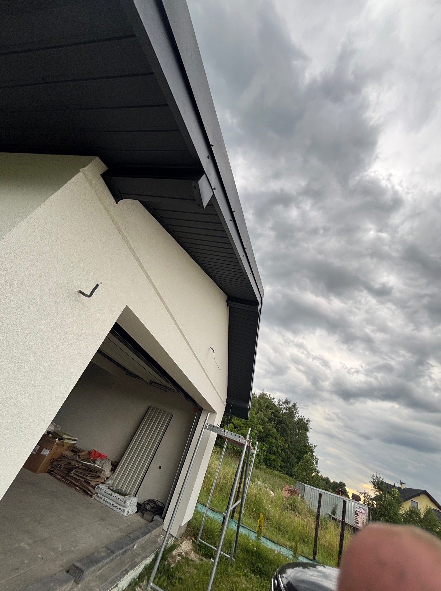 Nowoczesny dom z czarną podbitką dachową, widok z dołu na tle pochmurnego nieba. Częściowo otwarty garaż z materiałami budowlanymi w środku.