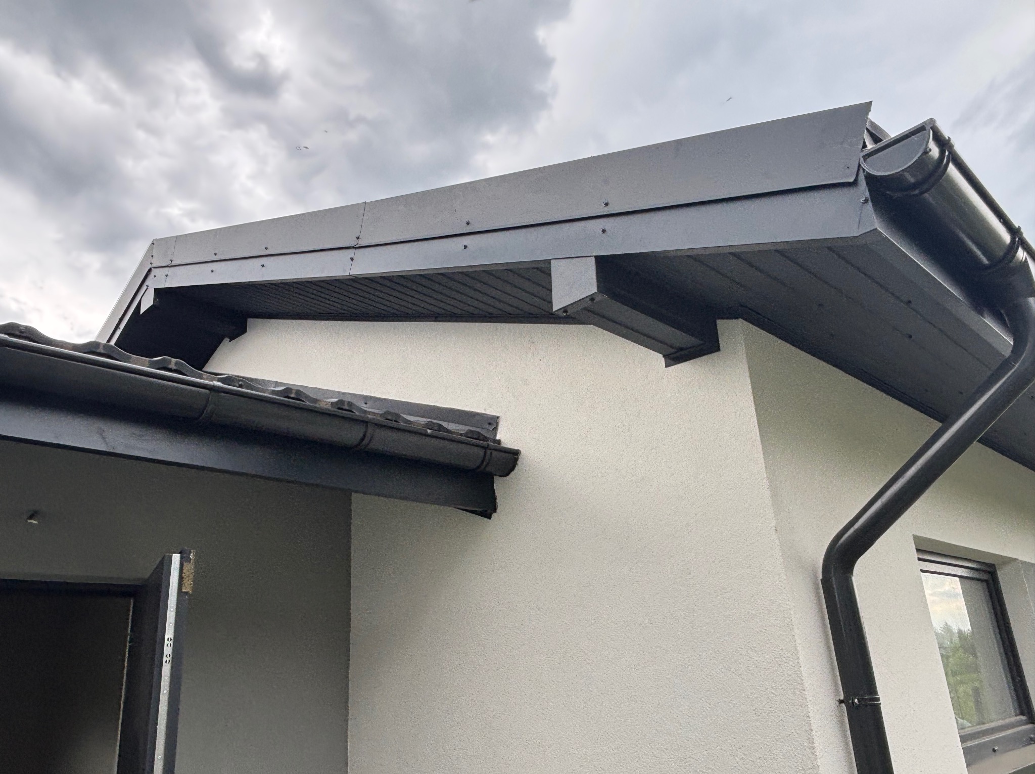 Detal czarnego dachu z rynnami i obróbkami blacharskimi na tle pochmurnego nieba. Nowoczesne wykończenie domu z widocznym fragmentem elewacji i okna.