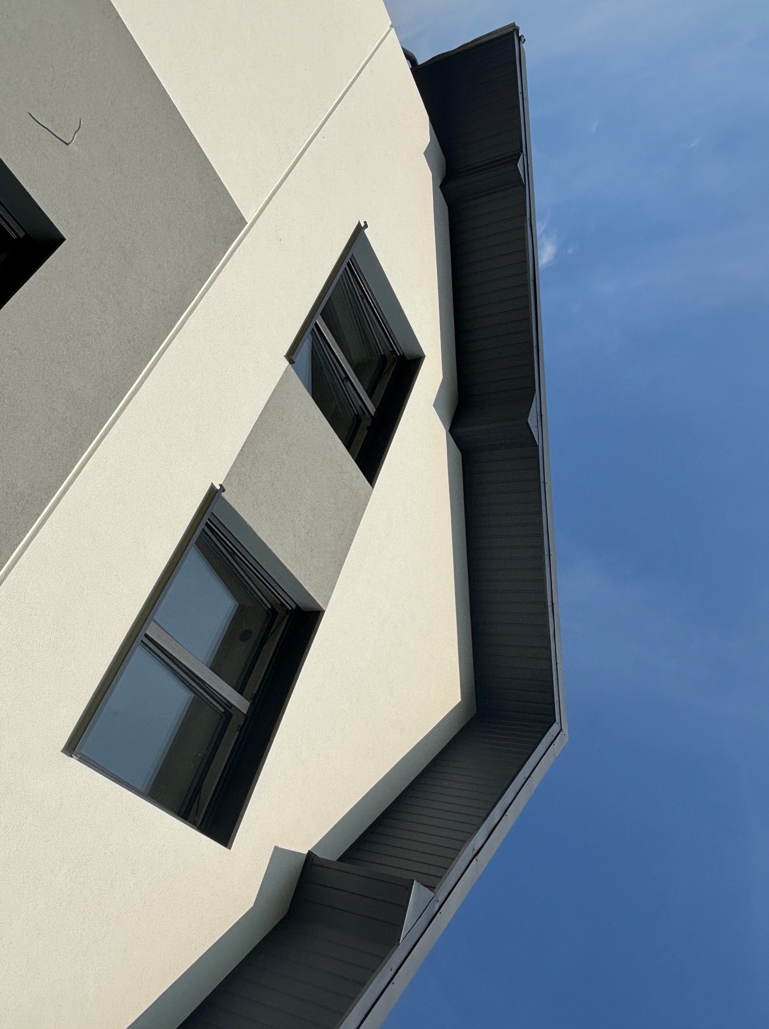 Nowoczesny budynek z szarym dachem i rynnami, widziany z dołu na tle błękitnego nieba. Minimalistyczna architektura z prostokątnymi oknami.