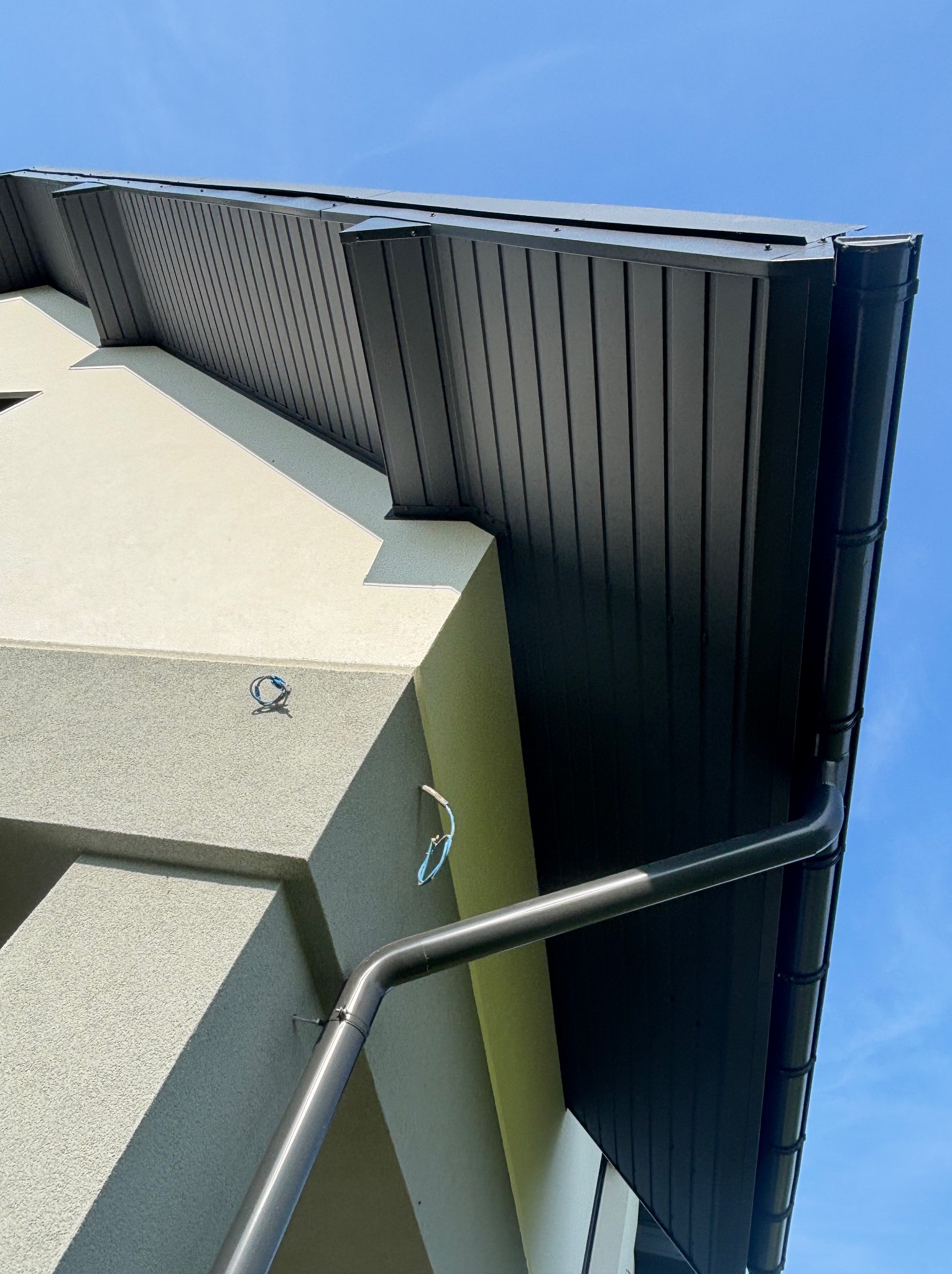 Elewacja domu z zamontowaną, czarną rynną i podbitką dachową. Widok z dołu na fragment budynku i błękitne niebo.