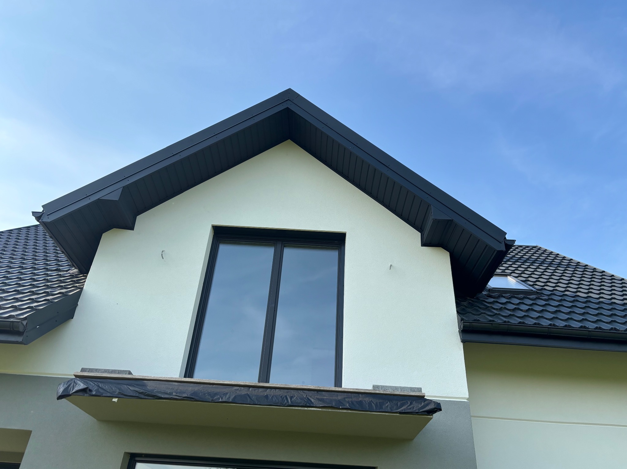 Fragment domu z czarną dachówką i obróbkami blacharskimi w kolorze antracytu. Widoczne okno dachowe i balkon z zabezpieczeniem przed zabrudzeniem.