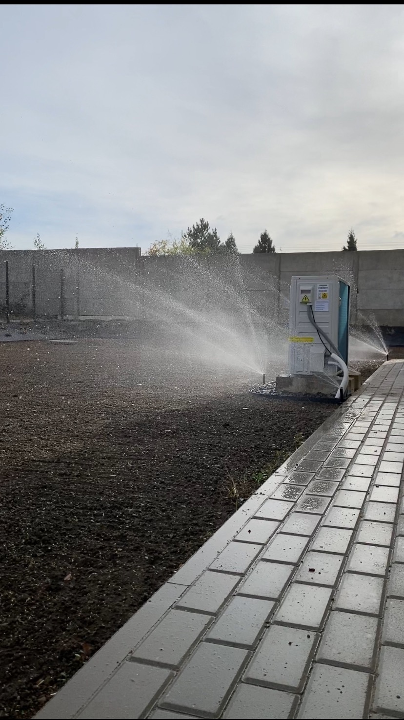 Automatyczny system nawadniania trawnika w trakcie pracy, widoczny zraszacz i woda, świeżo przygotowana ziemia pod trawnik, ułożona kostka brukowa.