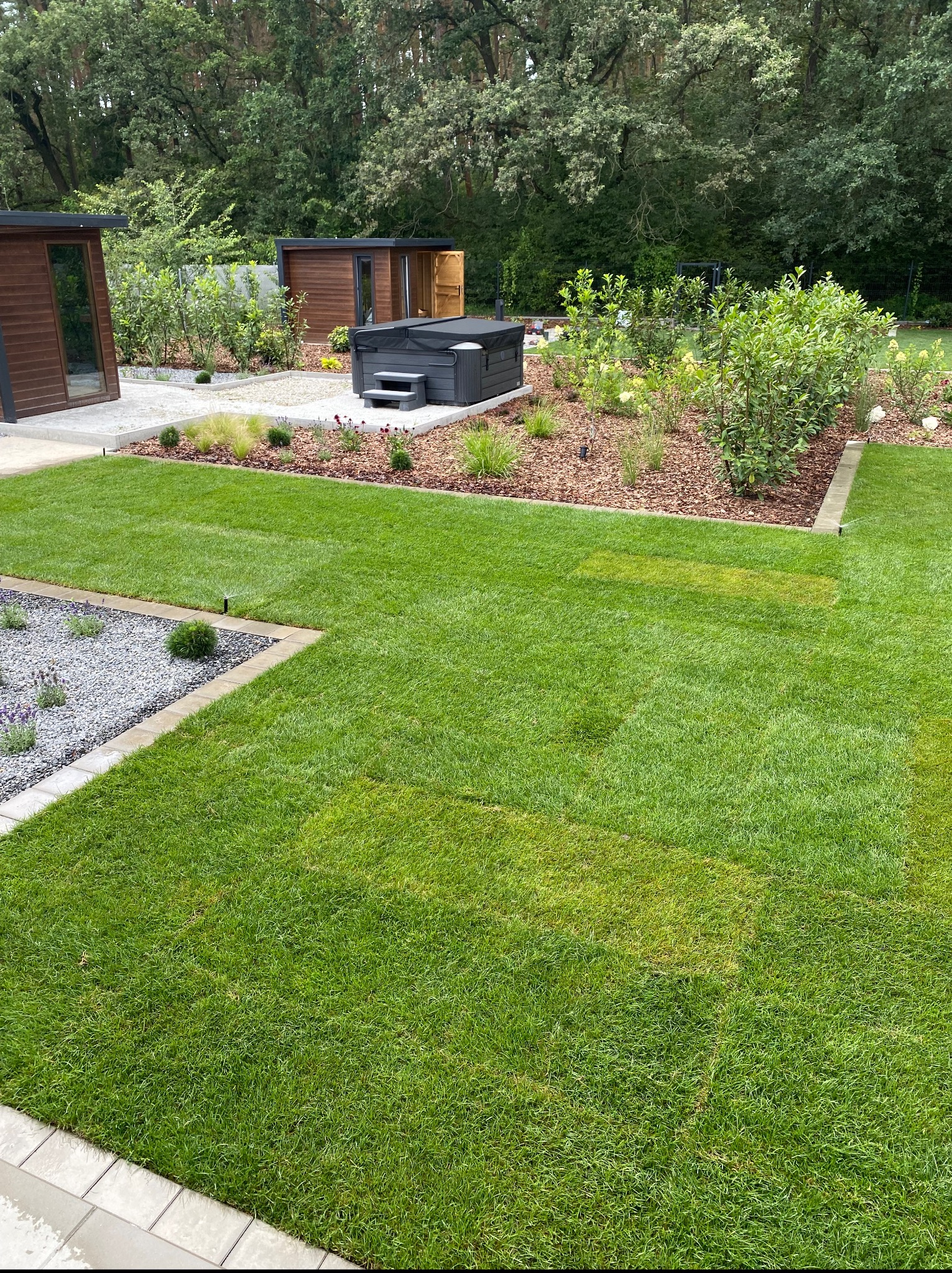 Zadbany ogród z równo przystrzyżonym trawnikiem, ozdobnymi rabatami z korą i kamieniami, oraz drewnianymi budynkami w tle, w słoneczny dzień.