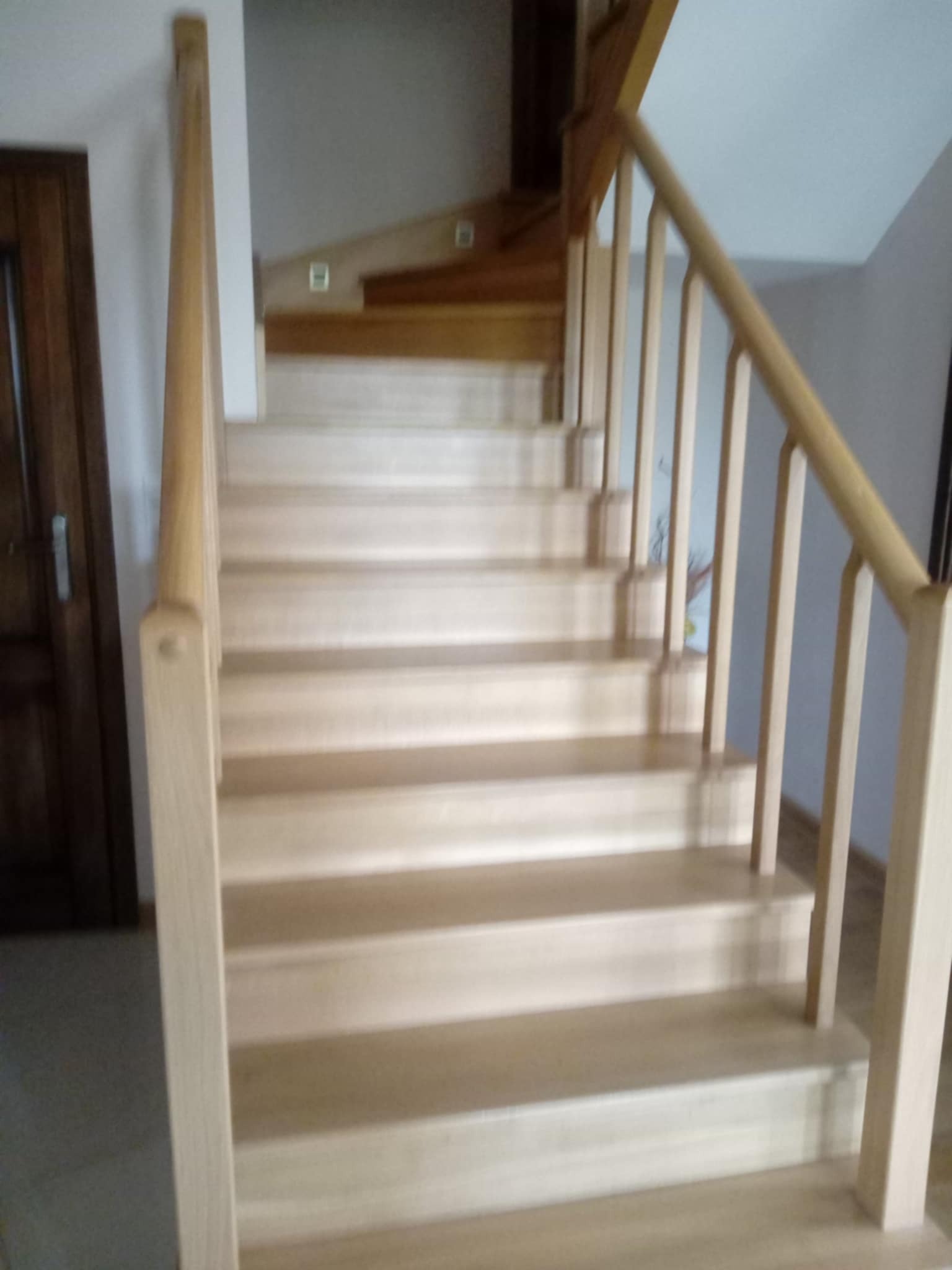 Jasne, drewniane schody z poręczą, widok z dołu. Stopnie schodów prowadzą w górę, w tle widoczne drzwi i fragment ściany.