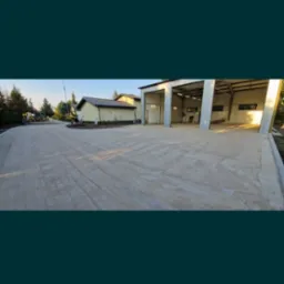 Nowa, betonowa nawierzchnia przed budynkiem z otwartymi wrotami garażowymi, widok z poziomu gruntu, maszyny budowlane w tle.