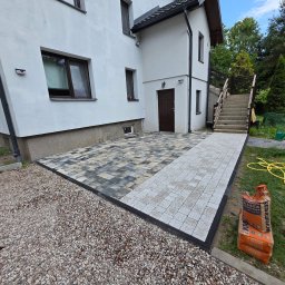 DYB-BUD MICHAŁ DYBOWSKI - Ułożona kostka brukowa w dwóch odcieniach szarości tworzy wzór przed budynkiem. Obrzeża z ciemnej kostki. Obok leżą worki z piaskiem kwarcowym.