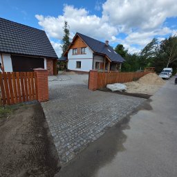 DYB-BUD MICHAŁ DYBOWSKI - Ułożona kostka brukowa na podjeździe posesji z domem i garażem, ogrodzenie z ceglanymi słupkami. Błękitne niebo z obłokami w tle.