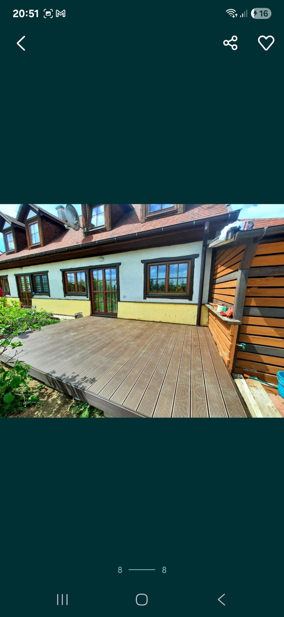 Drewniany taras przylegający do budynku z żółtą elewacją, widok z poziomu gruntu, zieleń w tle. Uwagę przykuwa brązowa kolorystyka desek tarasowych.