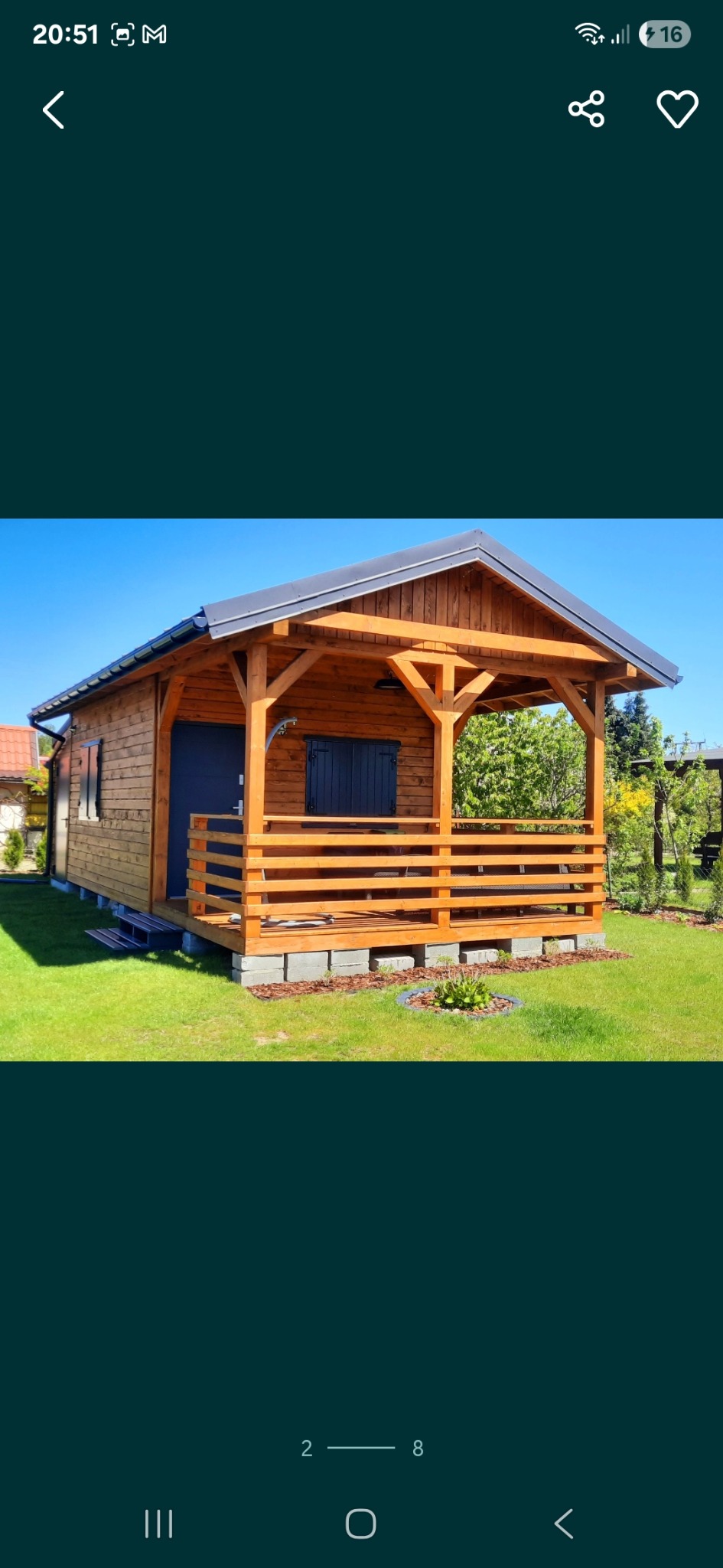 Drewniany domek letniskowy z zadaszonym tarasem, wykończony w naturalnym odcieniu drewna, na tle zielonego trawnika i błękitnego nieba. Widoczne detale wykończenia.