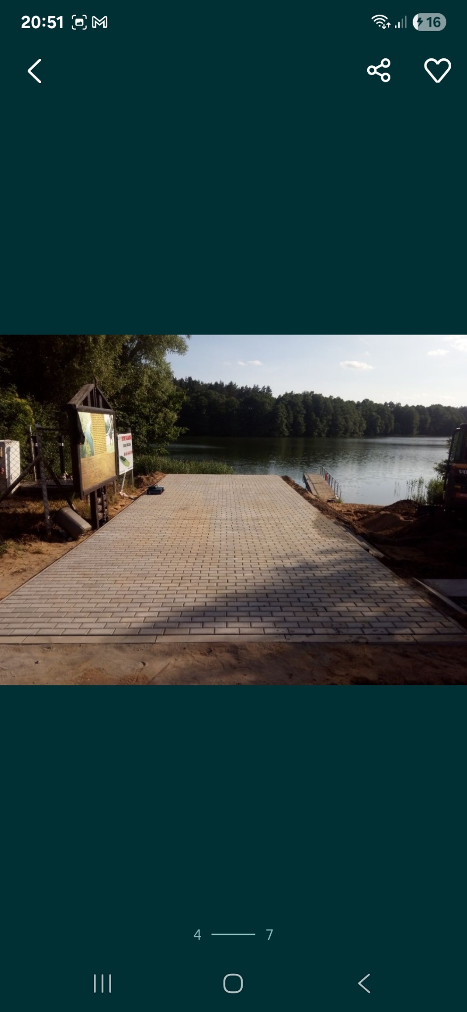 Wyłożona kostką brukową ścieżka prowadzi do jeziora, otoczona zielenią. Widoczny znak informacyjny i sprzęt budowlany. Kostka w odcieniach szarości, regularny wzór.