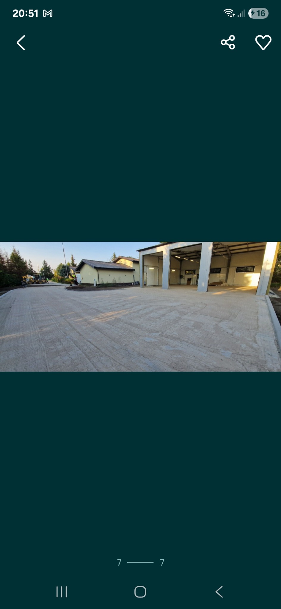 Nowa, betonowa nawierzchnia przed budynkiem z otwartymi wrotami garażowymi, widok z poziomu gruntu, maszyny budowlane w tle.