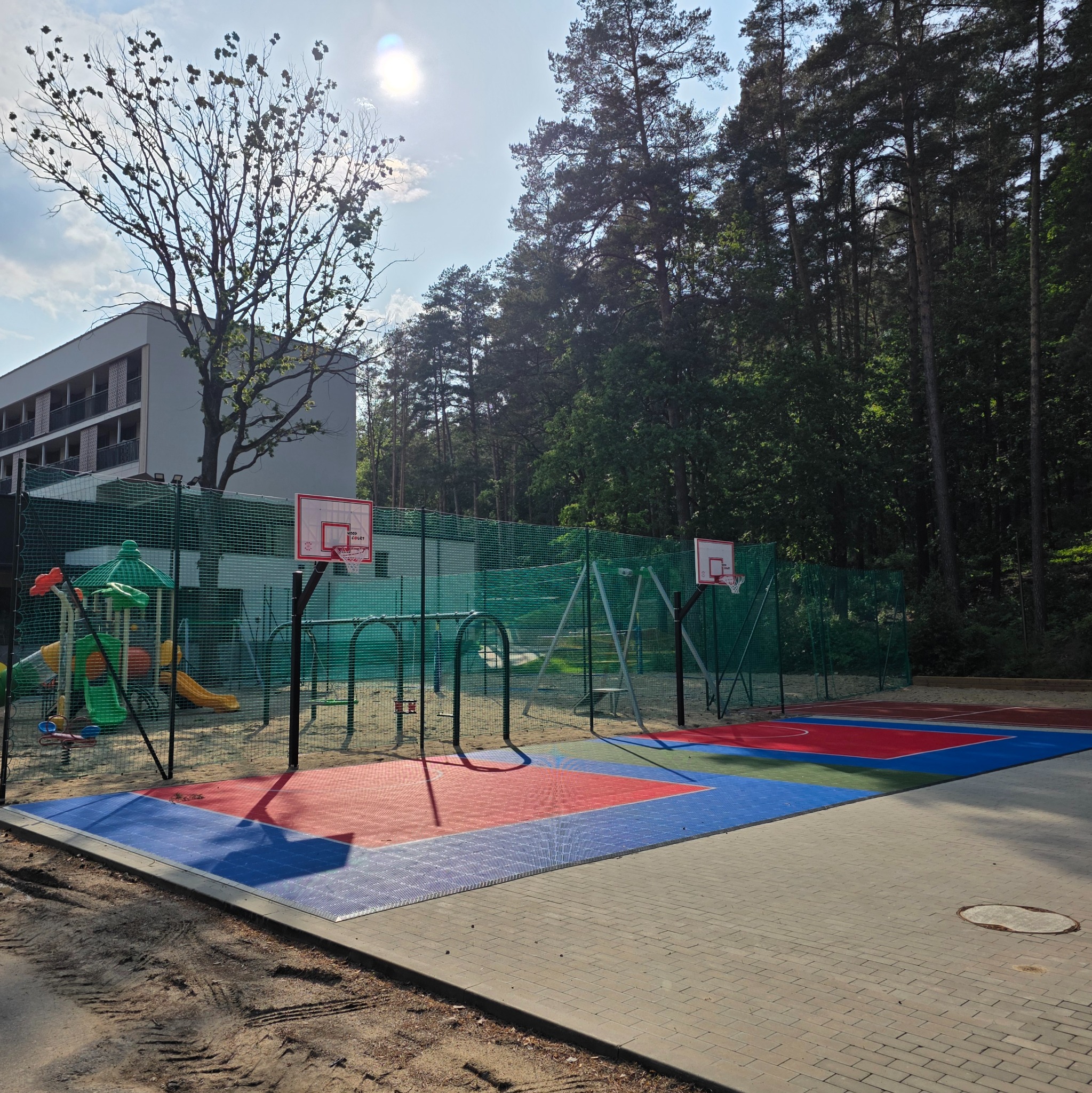 Kolorowe boisko z kostki brukowej i plac zabaw za siatką, otoczone drzewami i budynkiem w tle, w słoneczny dzień. Widoczne ślady po pracach ziemnych.