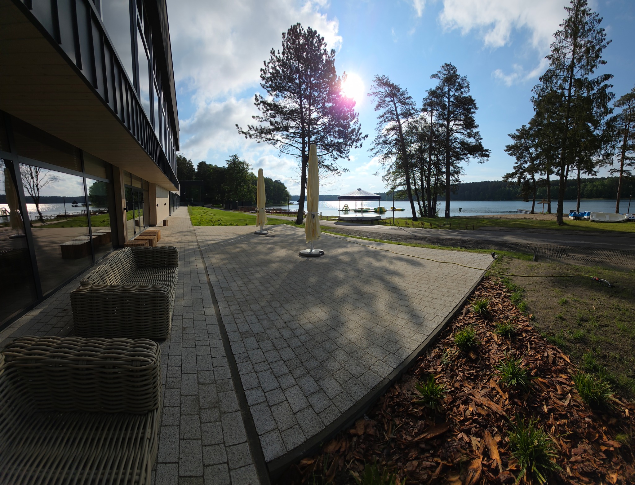 Kostka brukowa tworzy patio przy nowoczesnym budynku z widokiem na jezioro. Widoczne rattanowe meble ogrodowe i parasole. Słoneczny dzień, błękitne niebo.