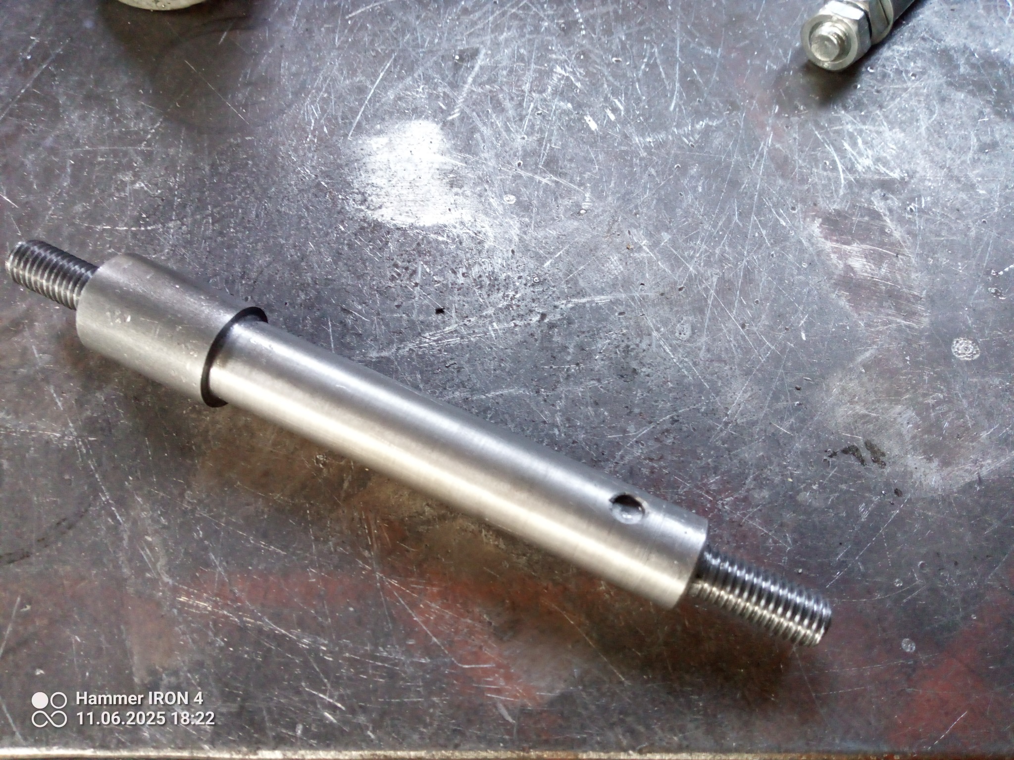 Precyzyjnie wykonany, gwintowany metalowy wałek z otworem, leżący na metalowej powierzchni z widocznymi śladami obróbki. Detal gotowy do montażu.