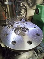 Obrabiany metalowy element na tokarce z widocznymi wiórami, oświetlony punktowo, z detalami maszyny w tle. Widok z góry na okrągły element.