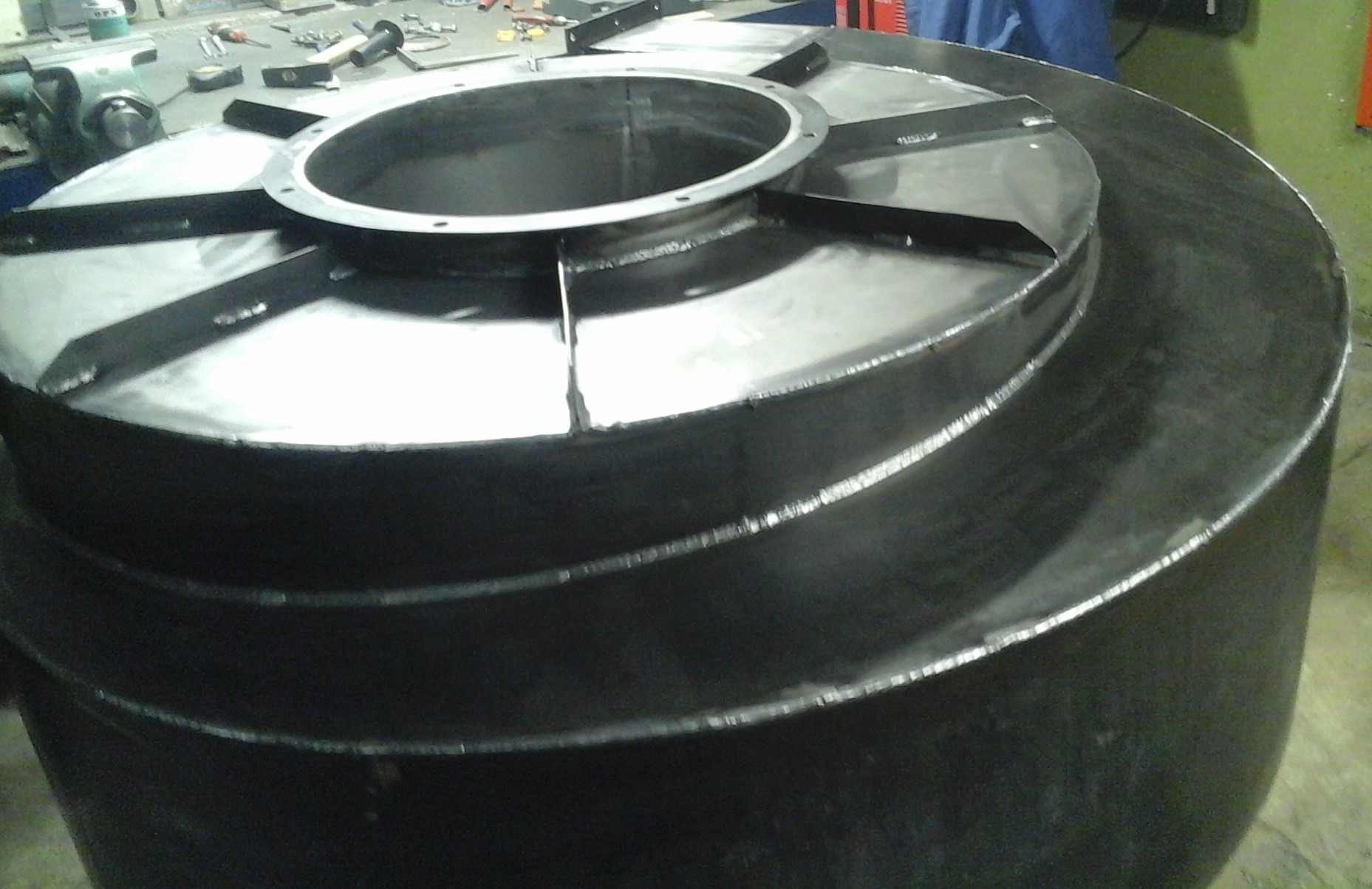 Metalowa konstrukcja zbiornika z widocznymi spawami i elementami mocującymi, wykonana z ciemnego metalu, w warsztacie.