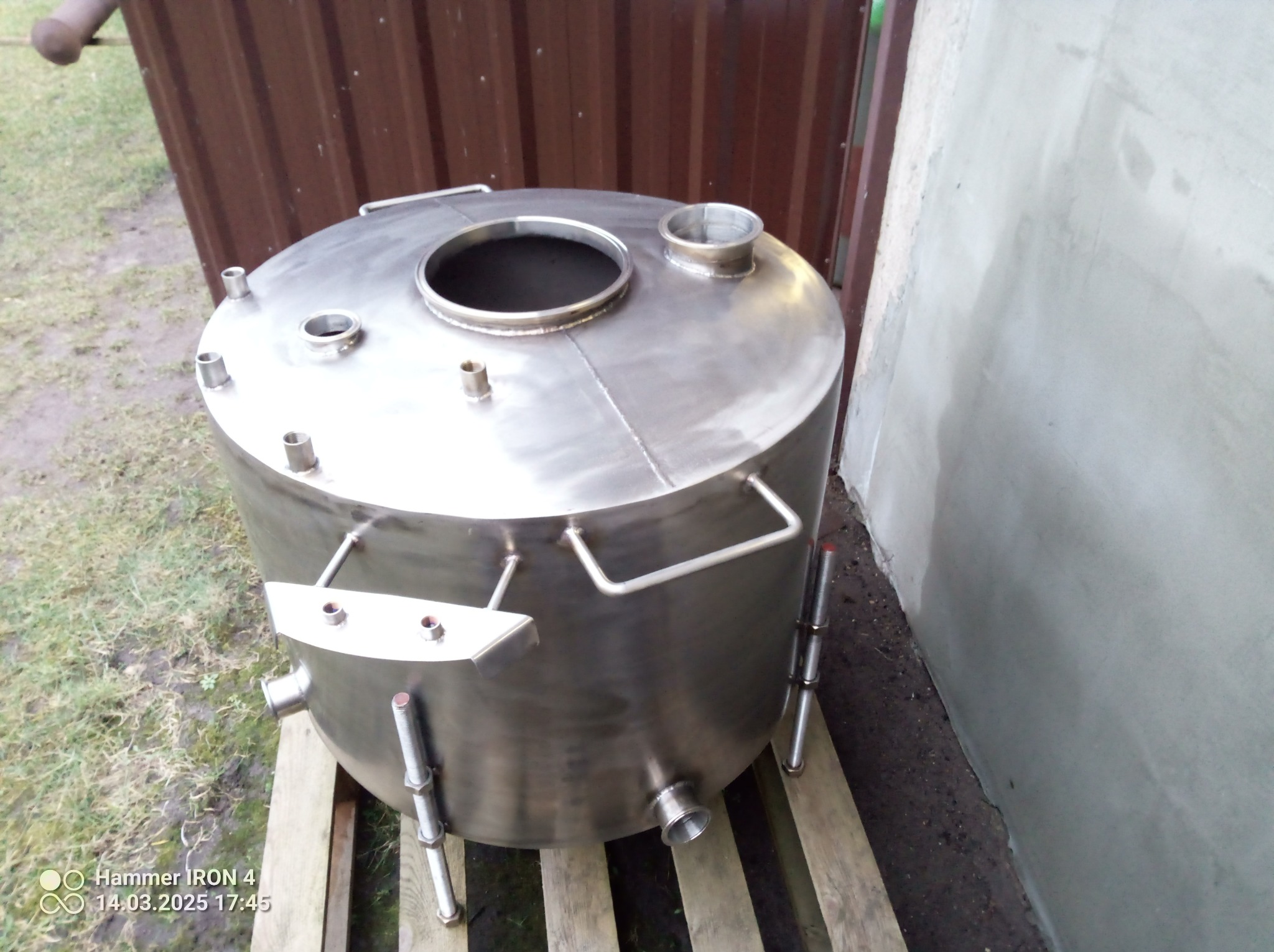 Metalowy zbiornik ze stali nierdzewnej na palecie, widoczne spawane łączenia, króćce i uchwyty. Tło stanowią brązowe drzwi i szara ściana.