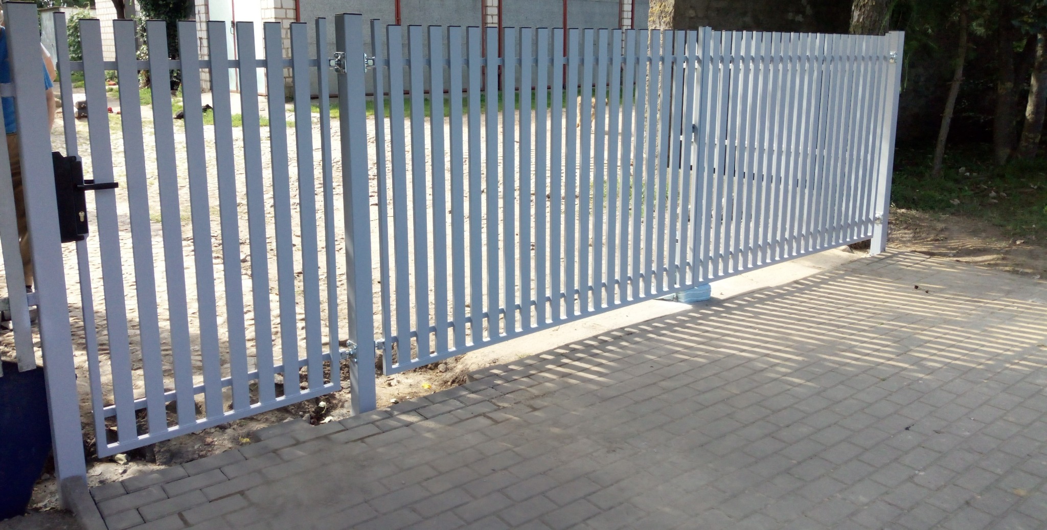 Szara, metalowa brama posesyjna z pionowymi szczeblami, częściowo otwarta, z widocznym zamkiem i kostką brukową przed nią. Widok z zewnątrz posesji.
