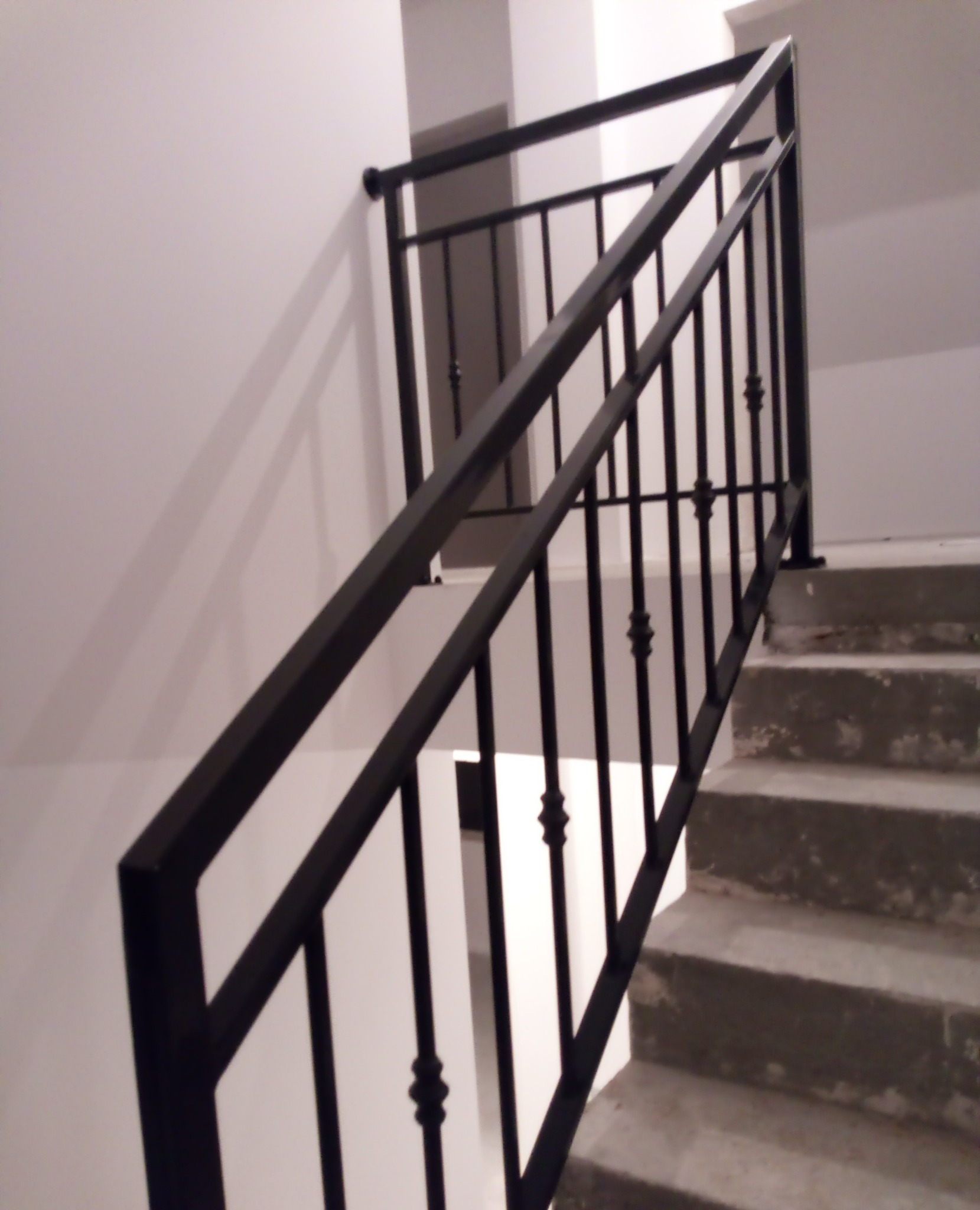 Nowoczesna, czarna balustrada schodowa z metalu, z dekoracyjnymi elementami, zamontowana przy betonowych schodach w jasnym wnętrzu. Ujęcie z dołu, podkreślające geometrię.