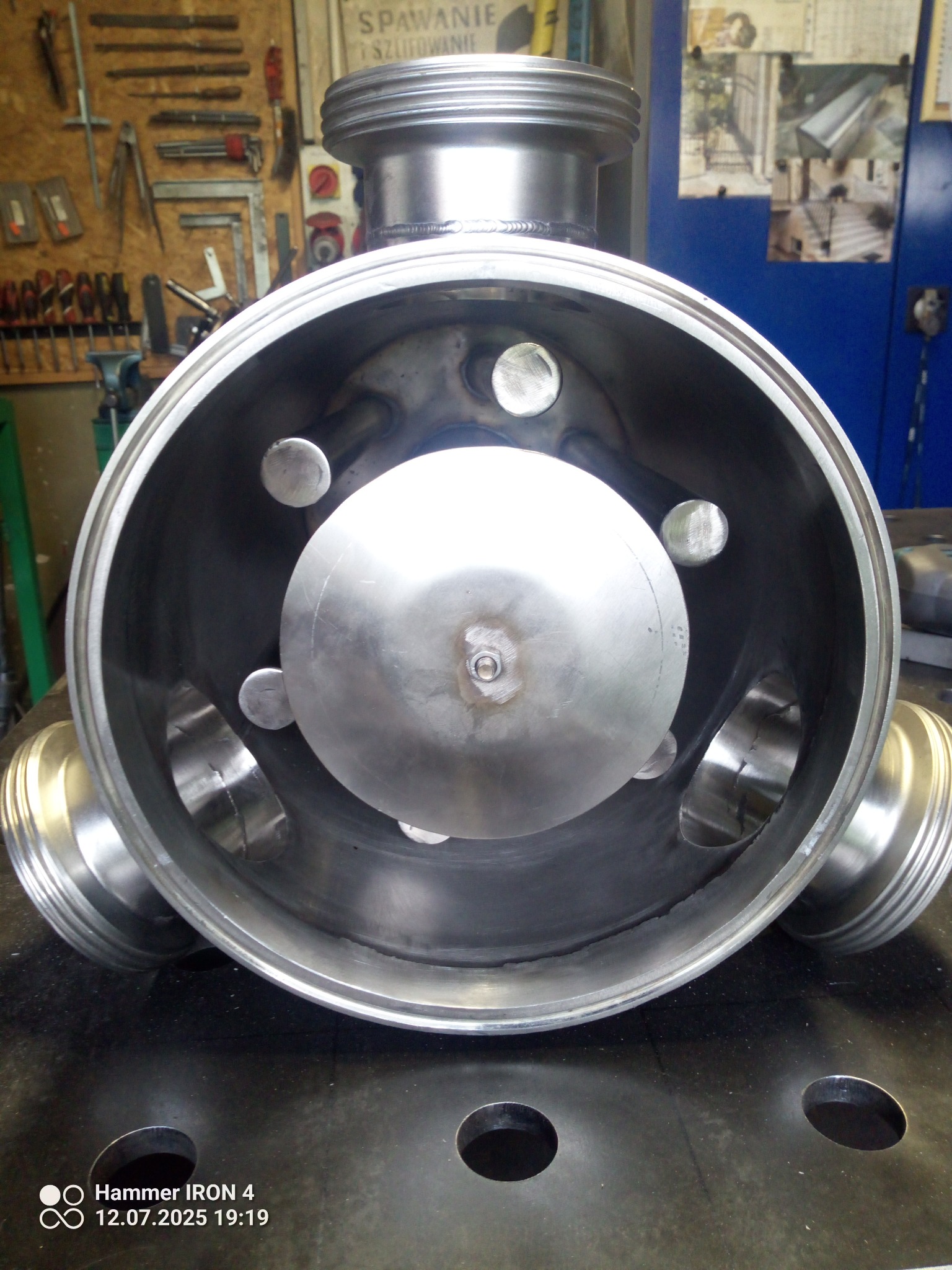 Metalowa konstrukcja cylindryczna z trzema otworami bocznymi i wewnętrznym elementem centralnym, widoczna na tle warsztatu. Precyzyjna obróbka metalu.