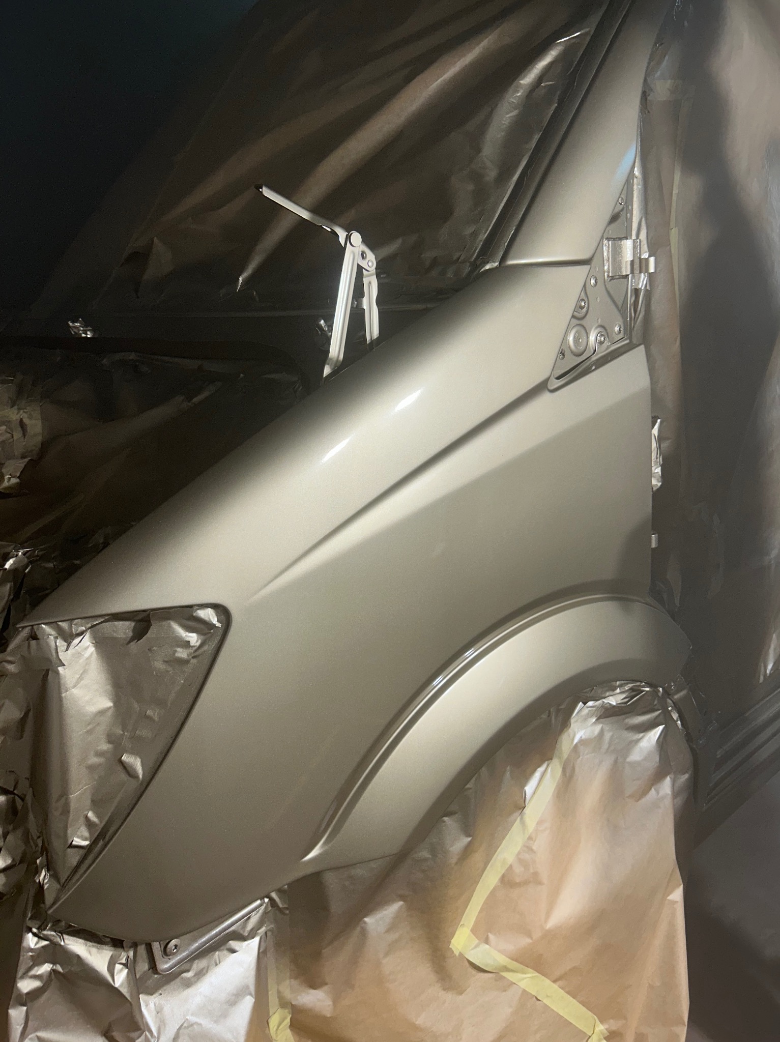 Lakierowany na złoto błotnik samochodu, zabezpieczony folią i papierem maskującym, w warsztacie w Kartuzach. Widoczne detale karoserii i świeża powłoka lakiernicza.