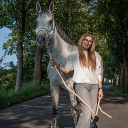 Kasia Krupa - Młoda kobieta w okularach, ubrana na szaro, prowadzi siwego konia po asfaltowej drodze w otoczeniu drzew.