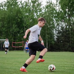 Kasia Krupa - Dynamiczne ujęcie piłkarza w akcji, skupionego na prowadzeniu piłki podczas meczu na zielonym boisku. W tle widoczni inni gracze i siatka bramki.