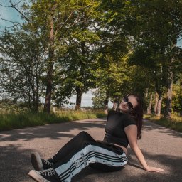 Kasia Krupa - Portret kobiety w okularach przeciwsłonecznych, siedzącej na asfaltowej drodze otoczonej drzewami, w sportowym stroju Adidas i trampkach Converse, z podpisem autora.