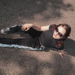 Kasia Krupa - Zdjęcie z góry: młoda kobieta w czarnym stroju sportowym i okularach przeciwsłonecznych leży na szarym chodniku. Widoczne buty typu Converse i cień osoby.