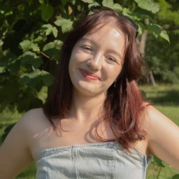 Kasia Krupa - Portret uśmiechniętej kobiety w plenerze, z zielonym tłem liści, w letniej stylizacji. Okulary na głowie i delikatna biżuteria dodają uroku.